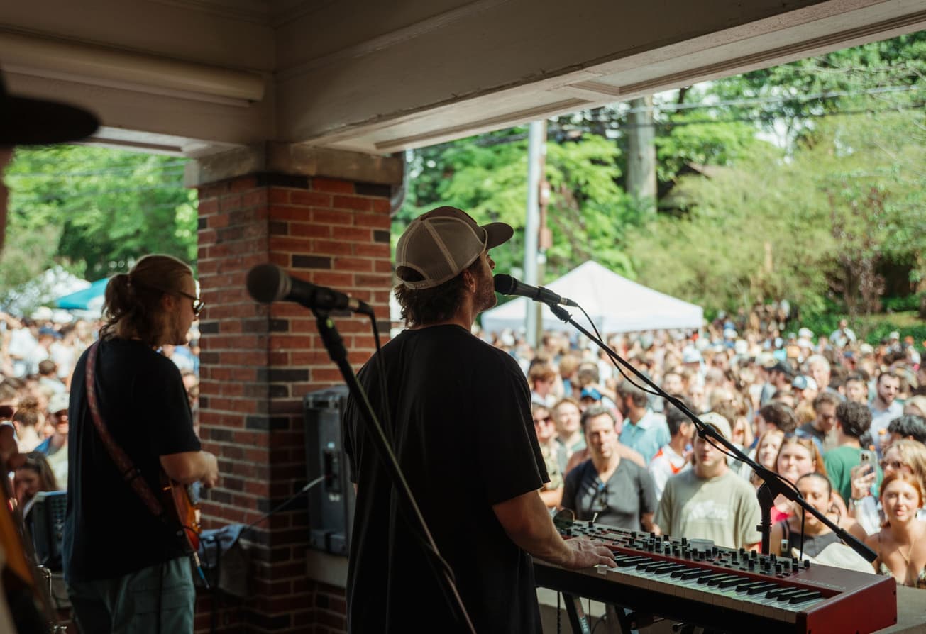 View from a porch stage: keyboardist and guitarist playing for a large outdoor crowd